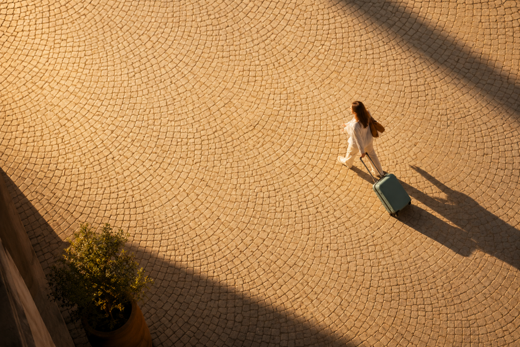 A traveler pulling luggage across a sunlit courtyard from above.